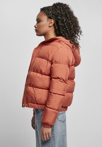 Pufferjakke i brent oransje, med hette, ribbestrikkede mansjetter og glidelås foran. Stoffet er skinnende med en quiltet tekstur.