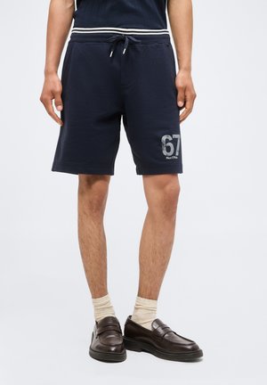 Personne portant un short bleu marine à cordon avec le numéro « 67 » imprimé sur la jambe gauche, associée à des chaussettes claires et des mocassins marron foncé.