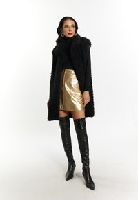 Vrouw in zwarte fuzzy mouwenloze jas, zwarte coltrui, metallic gouden mini-rok, zwarte kniehoge laarzen en gouden hoepel oorbellen.