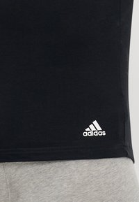 Juoda medvilnės marškinėliai su apvalia kaklo iškirpte, turintys baltą Adidas logotipą ant kairiojo apatinio krašto, derinami su šviesiai pilkais džinsais.