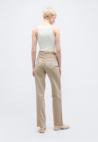 Άτομο με κοντά ξανθά μαλλιά, που φοράει λευκό αμάνικο τοπ, μπεζ τζιν με ψηλή μέση και μπεζ μπότες αστραγάλου με τακούνι, στέκεται μπροστά σε μονόχρωμο φόντο.