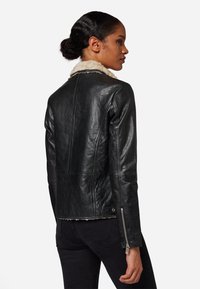 Zwarte leren jacket met een schapenvachtkraag, aangesloten ontwerp en ritssluitingen aan de mouwen. Textureerde oppervlakte met zichtbare stiksels en een rafelige zoom.