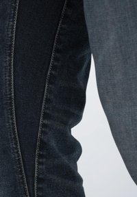 Jeans en denim noir avec des coutures contrastantes bleues, présentant une coupe ajustée et une texture visible. Le tissu affiche des variations de couleur subtiles.