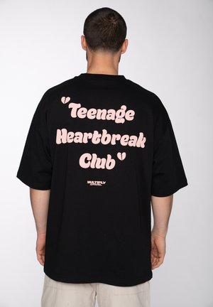 Mann mit kurzen Haaren, von hinten, trägt ein schwarzes oversized T-Shirt mit pinkem Text „Teenage Heartbreak Club“ und kleinem Markenlogo darunter.