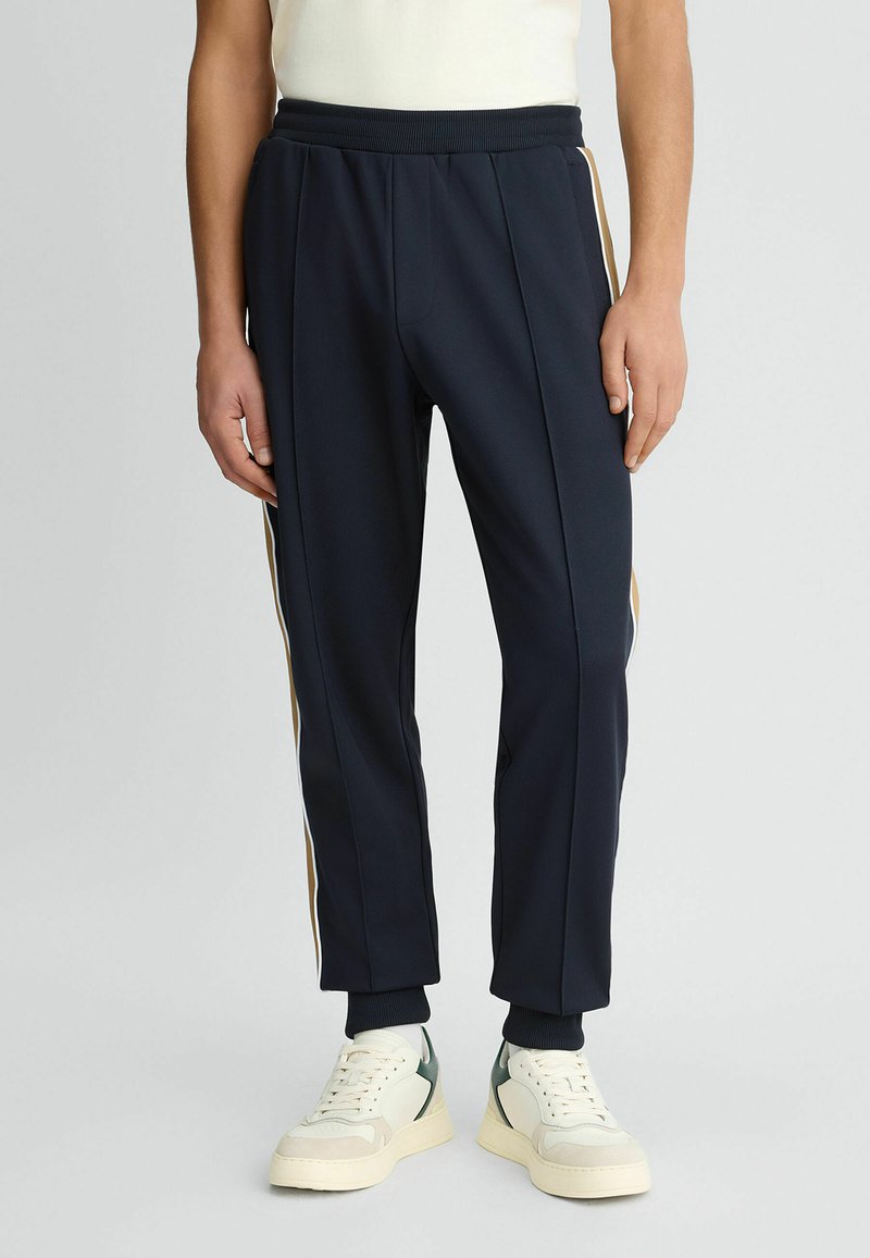 Marineblaue Sporthosen mit seitlichen goldenen und weißen Streifen, geripptem Bund und Bündchen, kombiniert mit hellen Sneakers. Glatte Stoffstruktur.