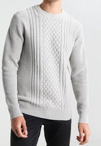 Man som bär en ljusgrå kabelstickad tröja med texturerade mönster och ribbade muddar, i kombination med svarta byxor.