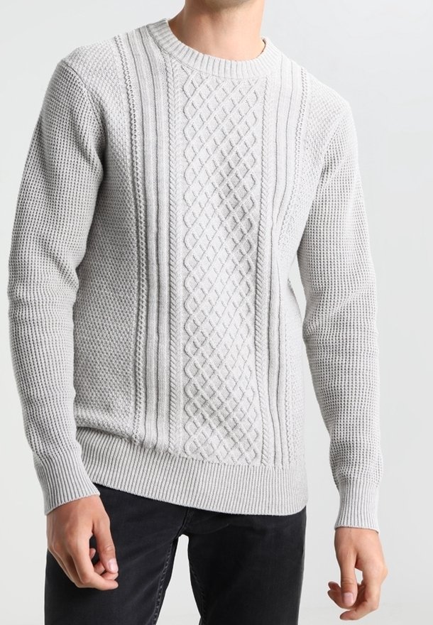 Man som bär en ljusgrå kabelstickad tröja med texturerade mönster och ribbade muddar, i kombination med svarta byxor.