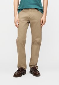 Άτομο που φοράει μπεζ παντελόνι ίσιας γραμμής, μπλε-πράσινο πουκάμισο περασμένο μέσα και σκούρα καφέ δερμάτινα δετά παπούτσια, στέκεται σε απλό ανοιχτό φόντο.