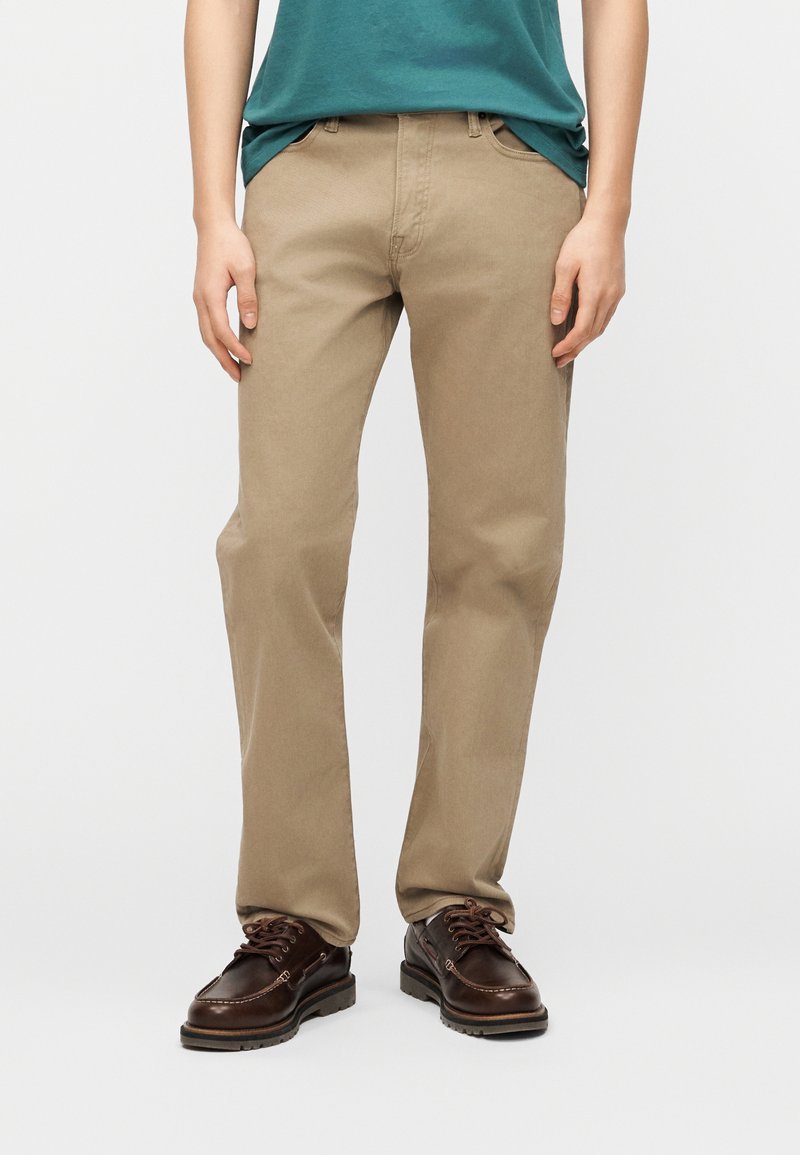 Άτομο που φοράει μπεζ παντελόνι ίσιας γραμμής, μπλε-πράσινο πουκάμισο περασμένο μέσα και σκούρα καφέ δερμάτινα δετά παπούτσια, στέκεται σε απλό ανοιχτό φόντο.