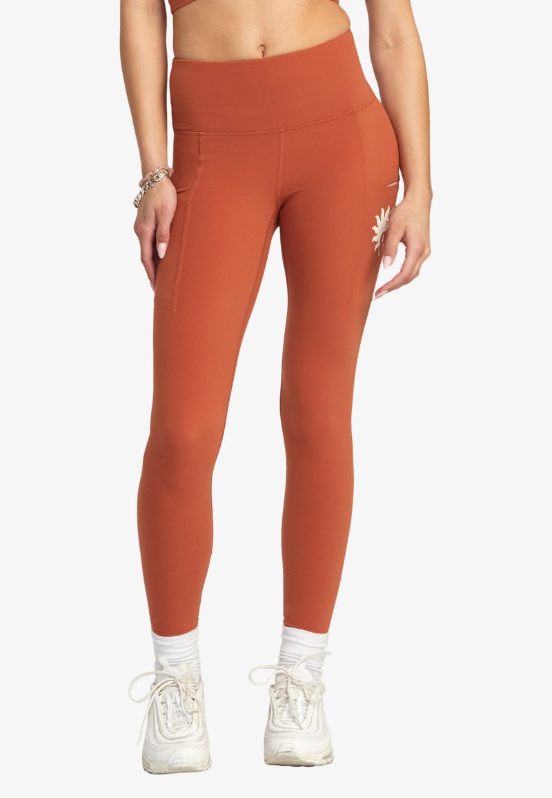 Rostfärgade leggings med hög midja, med en sidficka och ett solsymbol. Tyget verkar slätt och elastiskt.