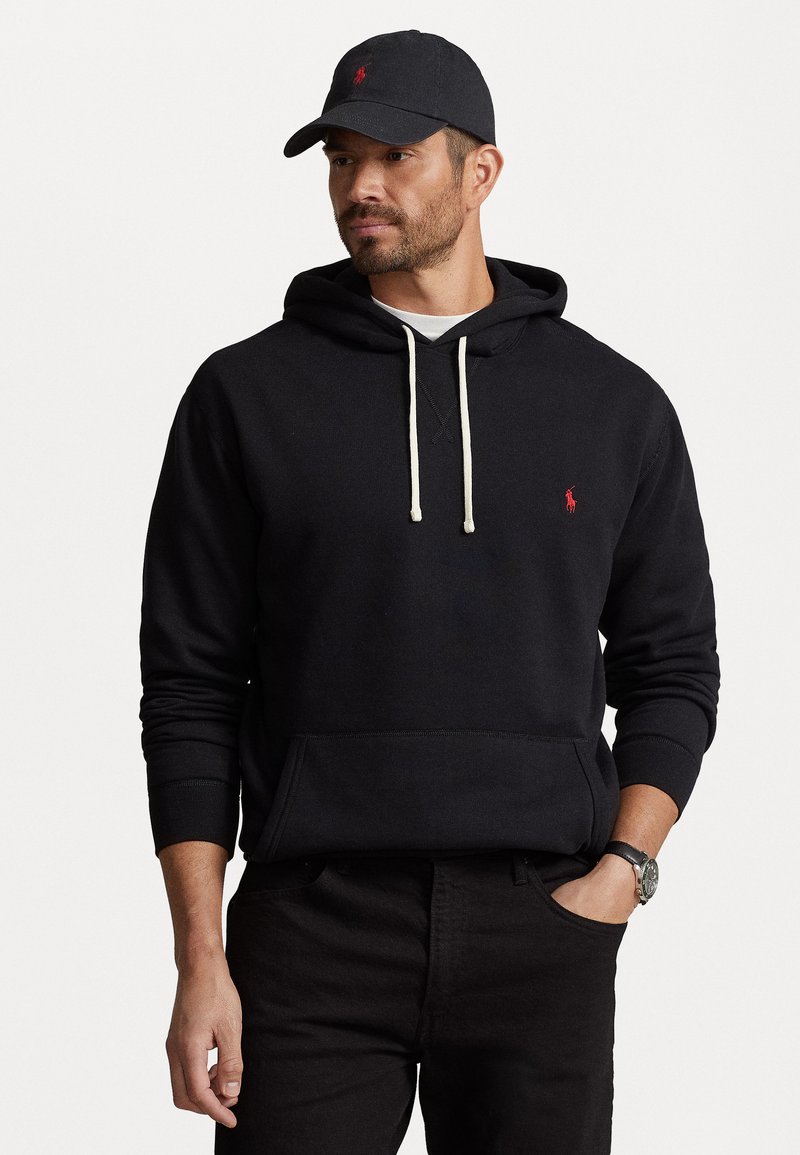 Črn bombažni shirt s kenguru žepom, kontrastnimi kremnimi vrvicami in majhnim rdečim logotipom na prsih, oblečen s črnimi hlačami in kapa.