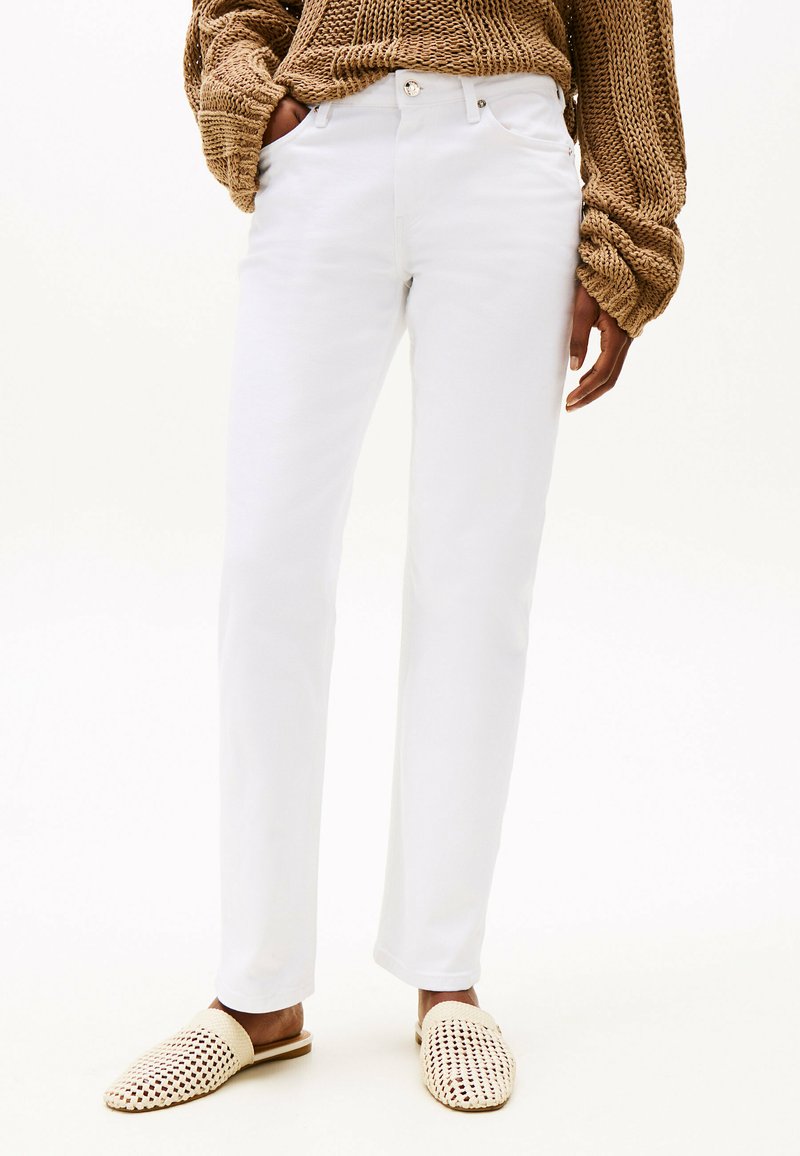 Pantalones blancos a medida con corte recto, diseño de cinco bolsillos y cierre de botón metálico, acompañados de zapatos de lona beige sin cordones.