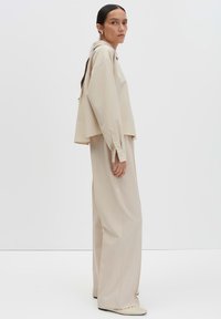 Donna con lunghi capelli scuri, in una camicia beige oversize, pantaloni larghi e scarpe piatte color crema, che sta in piedi di lato contro uno sfondo bianco.