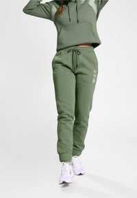 Grüne Kapuzenjacke mit einer Brusttasche und Kapuze, kombiniert mit passenden Jogginghosen mit elastischen Bündchen und Logo-Details. Weiße Sneakers runden das Outfit ab.