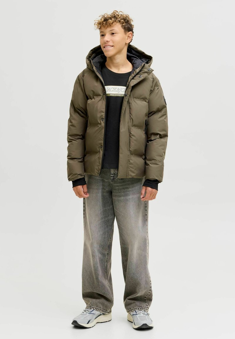 Olijfgroene pufferjack met capuchon, zwart t-shirt eronder, lichtgrijze wijde jeans en grijze sneakers met zwarte accenten.