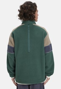 Fleece-Jacke in Grün, Beige und Marineblau. Merkmale sind ein hoher Kragen, ein halber Reißverschluss hinten und kontrastierende Nähte. Weicher Stoff mit oversized Passform.