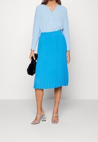 Top azul de manga larga, desabrochado en el pecho, combinado con una falda azul de talle rodilla con plisados. Bolso negro y sandalias de tacón bloque con estampado.
