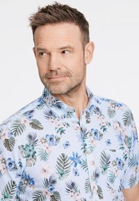 Camicia a maniche corte con bottoni di colore azzurro chiaro, decorata con un motivo floreale e foglie nei colori verde, blu e rosa, con una tasca sul petto.