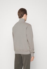 Felpa grigia con collo alto, polsini e orlo a costine, texture liscia e vestibilità relaxed, indossata sopra pantaloni scuri.