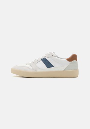 Sneakers bianche con striscia diagonale blu, dettagli in suede grigi e beige, materiali morbidi e texture consistenti, e suola color crema.