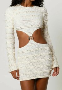 Cremfarbene Spitzen-Mini-Kleid mit langen Ärmeln und Rüschentextur, einem großen muschelförmigen Verschluss am Taillenausschnitt, getragen von einer Person mit dunkler Haut.