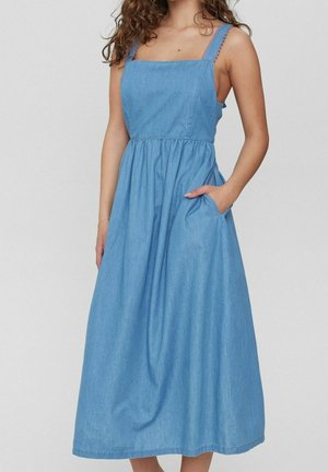 Femme portant une robe midi en denim bleu sans manches avec un décolleté carré et des poches, debout devant un fond uni clair.
