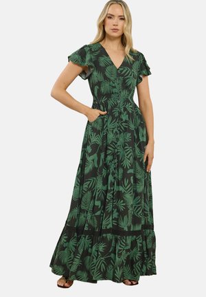 Vrouw draagt een zwarte maxi-jurk met groene tropische bladprint, korte wappermouwen, knopen aan de voorkant en een gerimpelde zoom, staande met één hand in de zak.