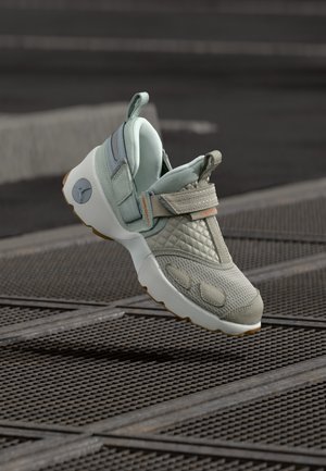 Chaussure de sport unique gris clair avec tissu matelassé et en maille, fermeture à bride et semelle blanche, exposée au-dessus d'une grille métallique en extérieur.