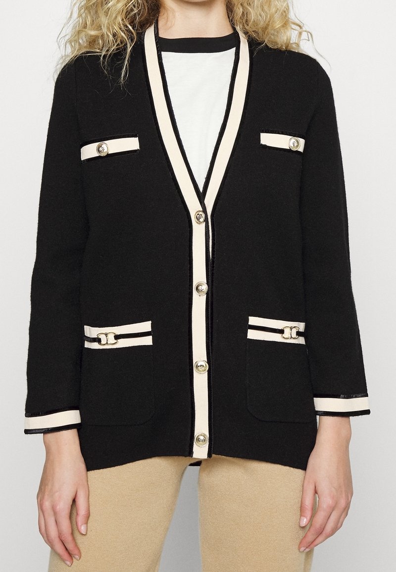 Femme portant un cardigan noir avec une bordure crème, des boutons dorés et des détails décoratifs aux poches, sur une chemise blanche et un pantalon beige.