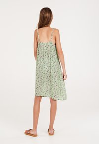 Groene jurk met bloemenpatroon, met dunne bandjes, losse pasvorm, halflange lengte en lichte stof. Draagt het met bruine sandalen.