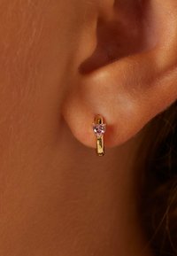 Gouden creool met een hartvormige roze edelsteen. Het ontwerp is minimalistisch met een gepolijste afwerking, tegen een huidkleurige achtergrond.