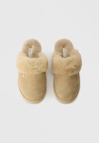 Tanfärgade mockasiner med en mjuk, lurvig kant, platta sulor och en liten metall-logo som accent. Har en rundad tå och en plyschoverdel.