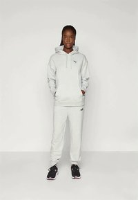 Giovane donna in piedi con le mani in tasca, indossando un hoodie grigio chiaro di Puma e pantaloni della tuta, e scarpe da ginnastica nere.