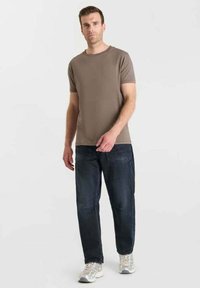 Uomo che indossa una maglietta marrone a maniche corte, jeans scuri e sneakers bianche, appoggiato a uno sfondo bianco uniforme.