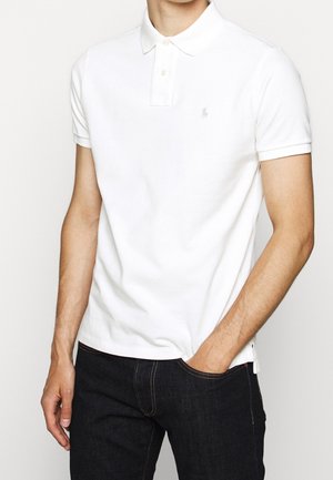 Mężczyzna ubrany w prostą białą koszulkę polo z krótkim rękawem, kołnierzykiem i trzema guzikami, w połączeniu z ciemnoniebieskimi dżinsami, z ręką w kieszeni.