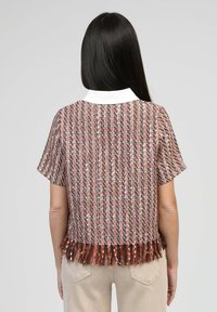 Femme aux longs cheveux bruns portant un haut texturé multicolore à manches courtes avec un col blanc et un ourlet à franges, un pantalon beiger, de dos.