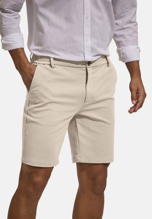 Mann trägt beige knielange Shorts mit Knopf und elastischem Bund, kombiniert mit einem weißen Langarmhemd mit hochgekrempelten Ärmeln.