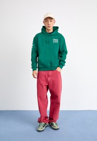 Green hoodie with a front pocket, white graphics. Loose-fitting red pants. Footwear: green sneakers with black accents. Neutral background.