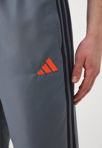 Graue Sporthosen mit schwarzen Streifen, ausgestattet mit einem leuchtend orangefarbenen gestickten Logo auf dem linken Bein. Glatte Textur und taillierte Passform.