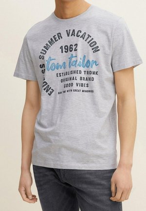 Mand iført lysegrå t-shirt med sort og blå tekst "Endless summer vacation 1962 tom tailor good vibes" og mørkegrå jeans.