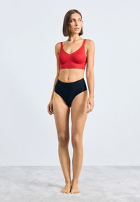 Vörös, textúrált bralette, hullámos széllel, magas derekú fekete bugyival párosítva. Mindkét darab sima felületű. Semleges háttér.