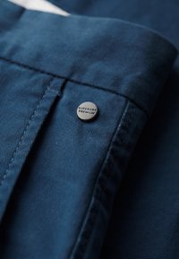 Tissu bleu foncé avec une texture subtile, présentant un bouton noir visible marqué "SUPERDRY PREMIUM" et des détails de couture le long de la couture.