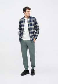 Καρό πουκάμισο φανέλας σε ναυτικό μπλε και κρεμ, φορεμένο πάνω από ένα λευκό μπλουζάκι. Απαλά πράσινα παντελόνια φόρμας και μαύρα αθλητικά παπούτσια ολοκληρώνουν το σύνολο.