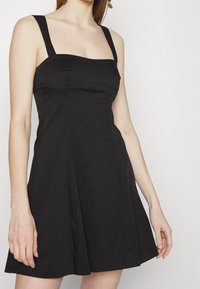 Svart ärmlös klänning med fyrkantig halsringning, figursydd överdel och utsvängd kjol. Tyget verkar slätt utan synliga mönster eller strukturer.