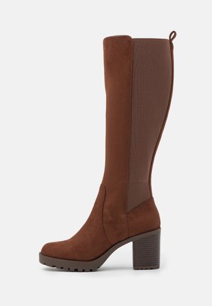 Bottes en cuir marron jusqu'au genou, en suède et en matière extensible, avec un bout arrondi, un talon épais et une semelle texturée.