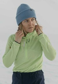 Haut à manches longues vert clair et texturé avec un col zippé quart, accompagné d'un bonnet en tricot bleu clair. Un pantalon foncé complète la tenue.
