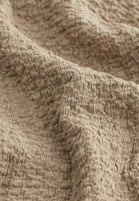 Beige strukturerad tyg med ett rynkigt mönster som visar varierande djup och en mjuk, följsam kvalitet. Slät yta med ett naturligt utseende.