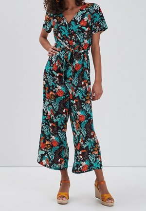 Femme portant une combinaison noire avec un motif floral coloré et des oiseaux tropicaux, associée à des sandales compensées jaune moutarde.