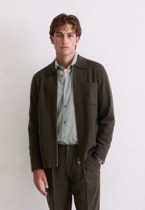 Veste zippée en laine marron avec col chemise, poche avant, associée à une chemise à carreaux verte et un pantalon marron assorti. Texture lisse.