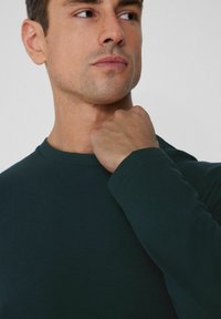 Ciemnozielona koszulka z długim rękawem i okrągłym dekoltem, wykonana z miękkiego materiału z dopasowanymi mankietami, prezentowana na modelu trzymającym kołnierz.